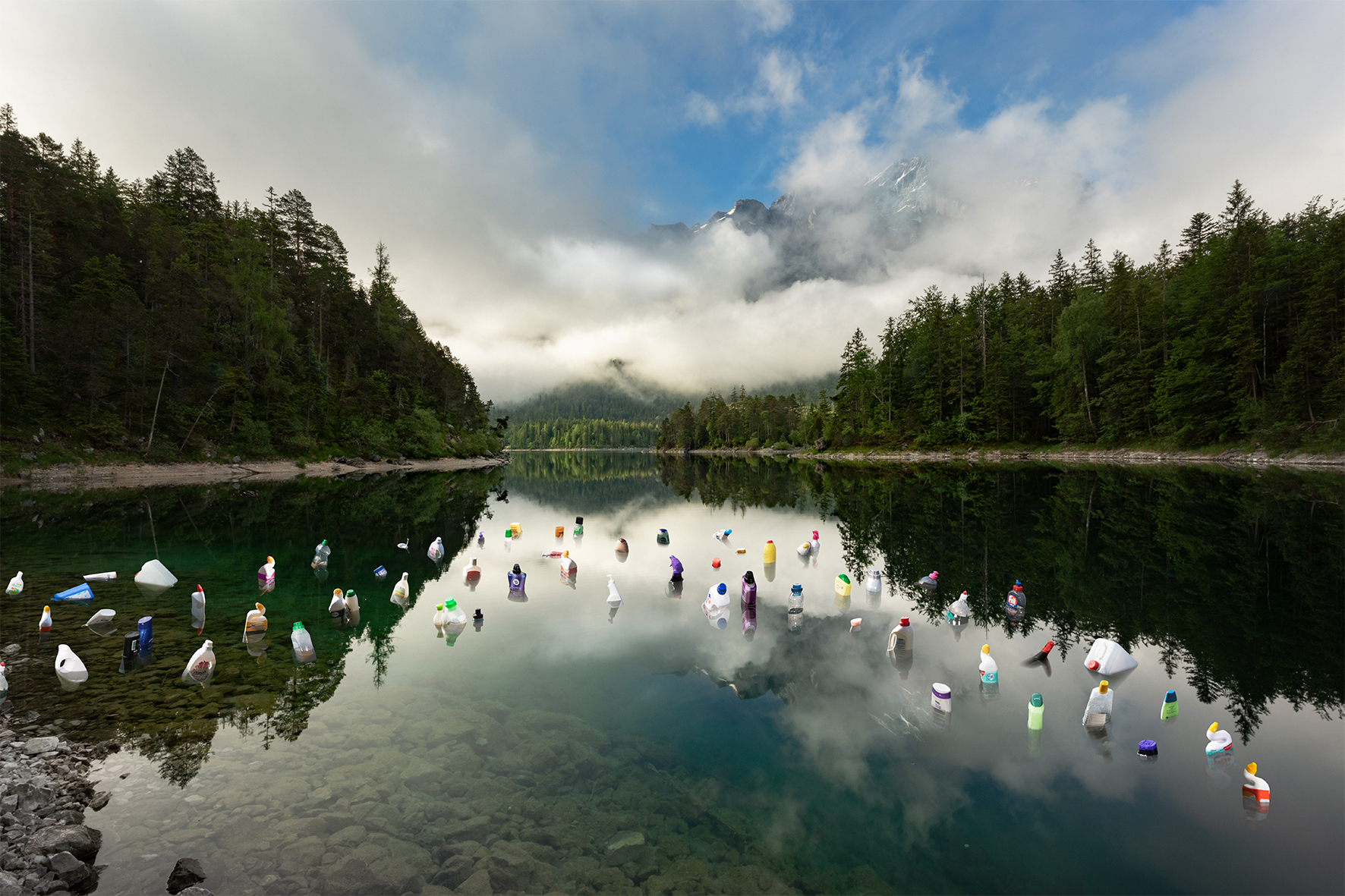 Kunstwerk von Dirk Krüll mit dem Namen Seepullen aus der Ausstellung Plastic Army, Ein See umringt mit idyllischer Landschaft und in dem See schwimmen leere Flaschen von Reinigungsmitteln