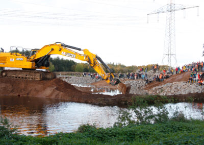 Bagger entfernt den Wall, Zuschauer stehen am Ufer